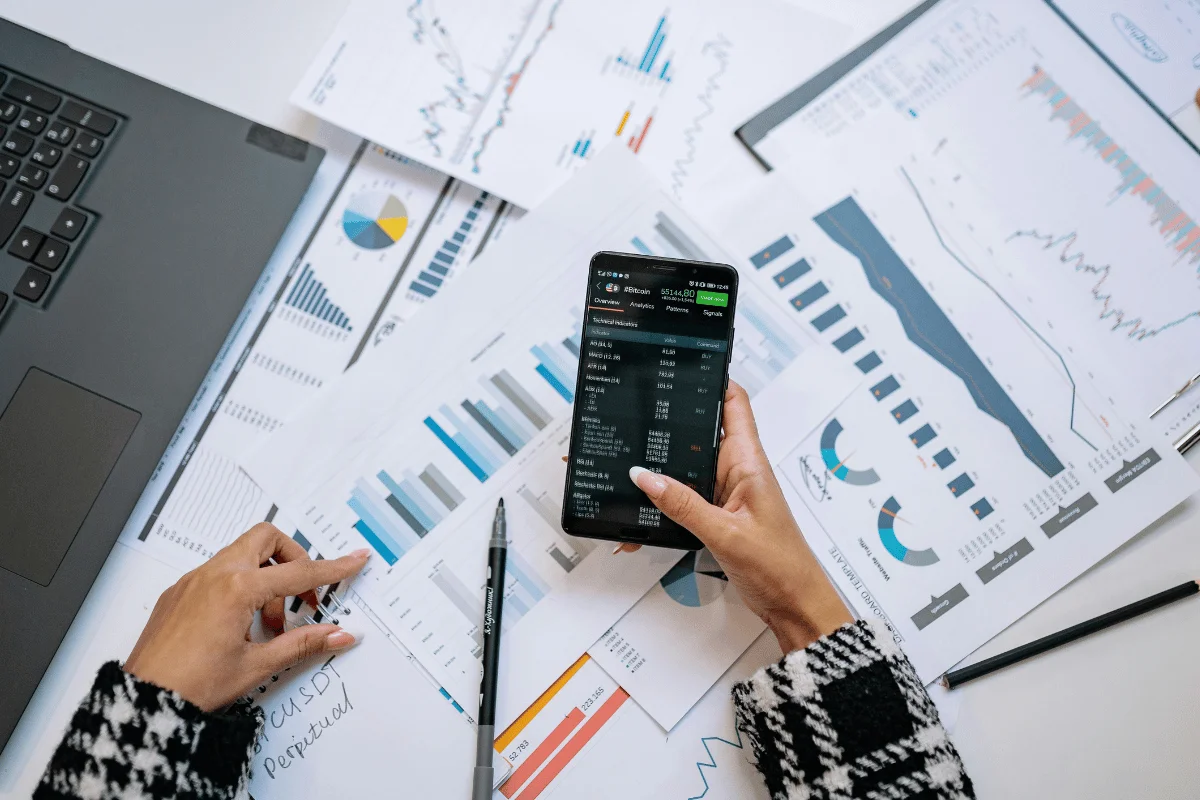 Hands holding a smartphone displaying financial data, surrounded by charts and graphs on paper, with a laptop nearby. Business and analysis theme.