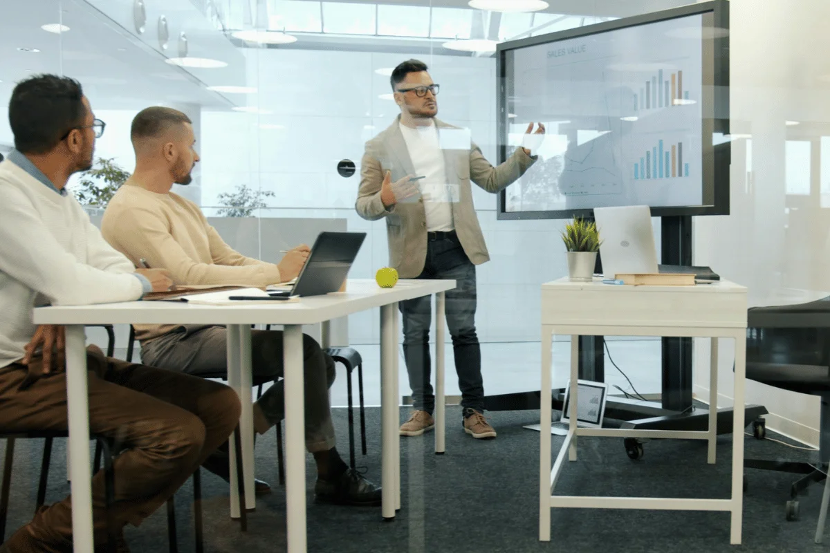 Business presenter explaining sales and performance charts to a team during a meeting in a modern glass office.