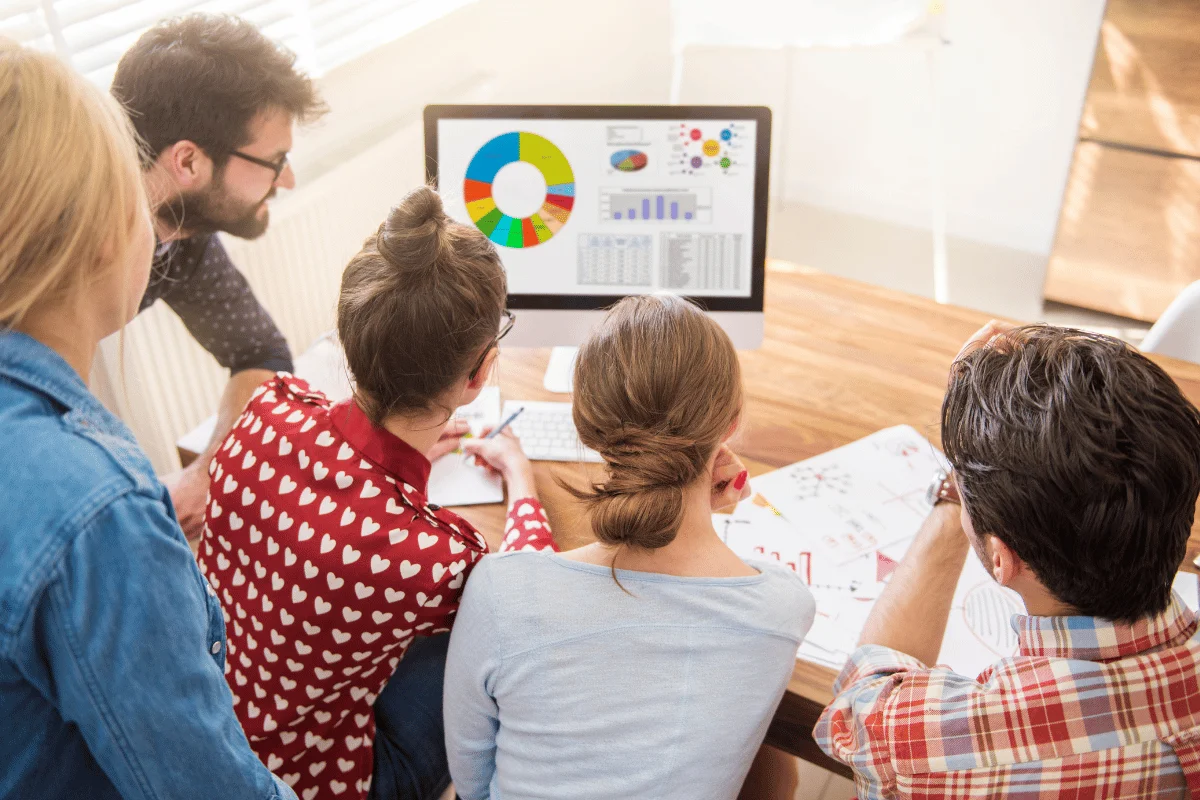 A group of five people looking at a computer screen displaying colorful charts and graphs. They appear engaged and focused, seated around a wooden table.