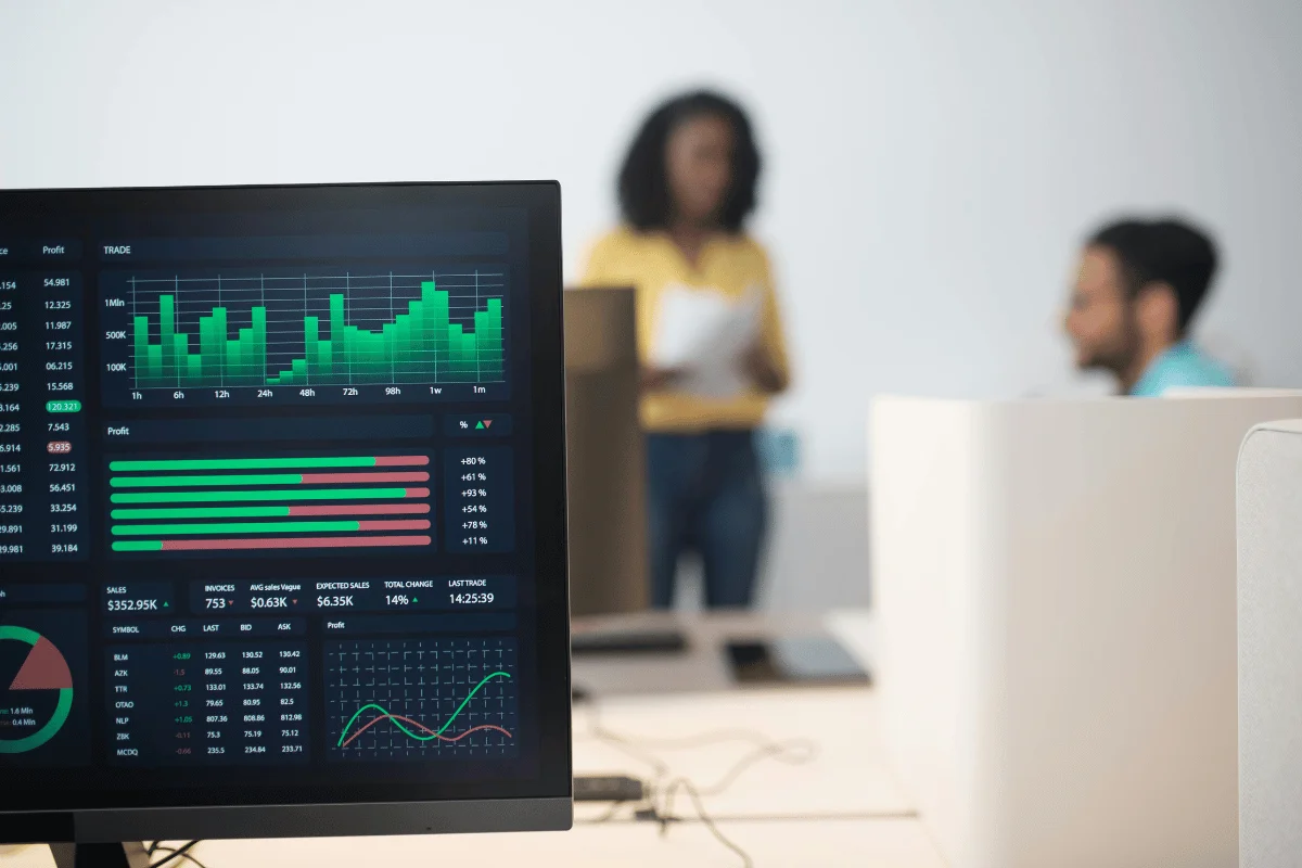 A computer screen displays financial graphs and data, focusing on bar and line charts. In the blurred background, two people engage in discussion.