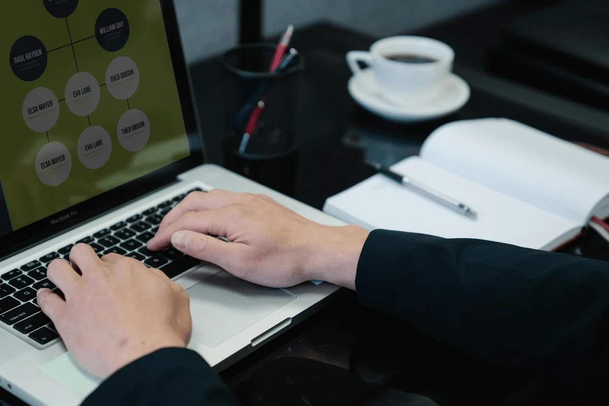 Hands typing on a laptop displaying a flowchart. Nearby, an open notebook with a pen and a cup of coffee on a desk, suggesting a focused work atmosphere.