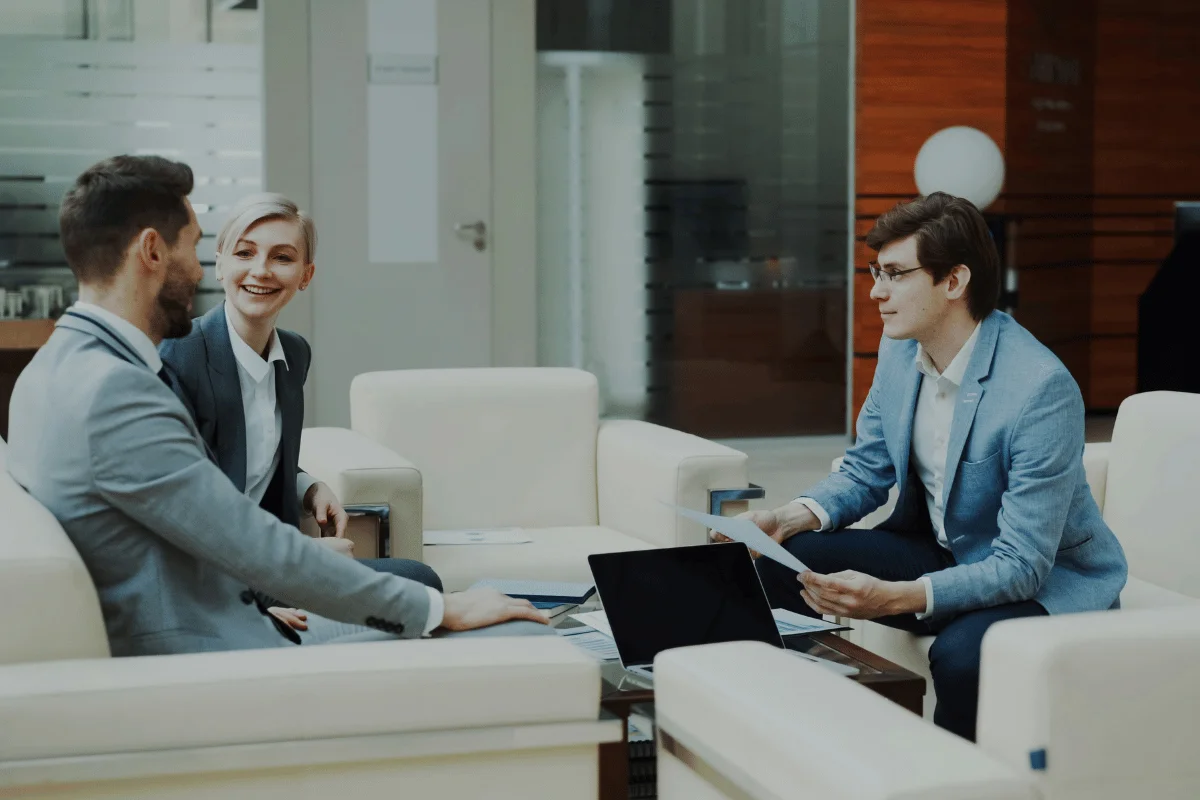 Three people in business attire converse in a modern office lounge. Two sit on a sofa, smiling, while the third, seated opposite, holds a document, fostering a collaborative atmosphere.