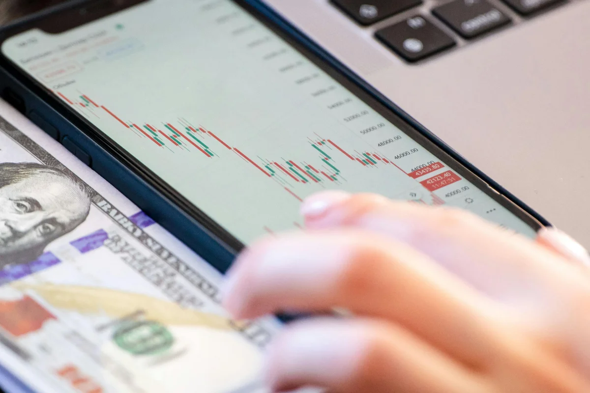 A hand interacts with a smartphone showing a stock chart, placed on a dollar bill and near a laptop keyboard.