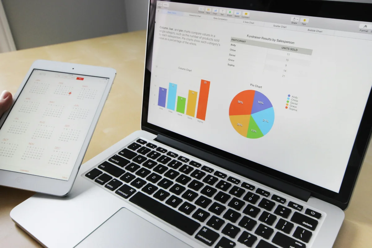 A person's hand holds a tablet displaying a calendar, while a laptop shows colorful charts analyzing fundraising results.