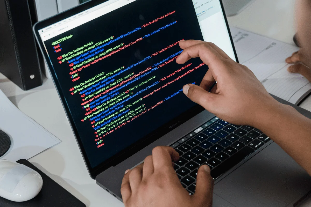 A person types code on a laptop screen displaying colorful HTML and JavaScript, on a desk with a notebook and a mouse nearby.