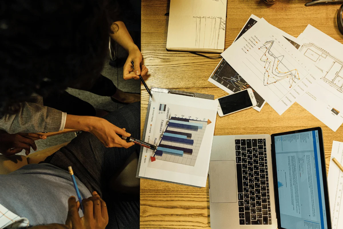 Top-down view of a conference table with diverse professionals analyzing financial bar charts. Includes a laptop, smartphone, and scattered documents.