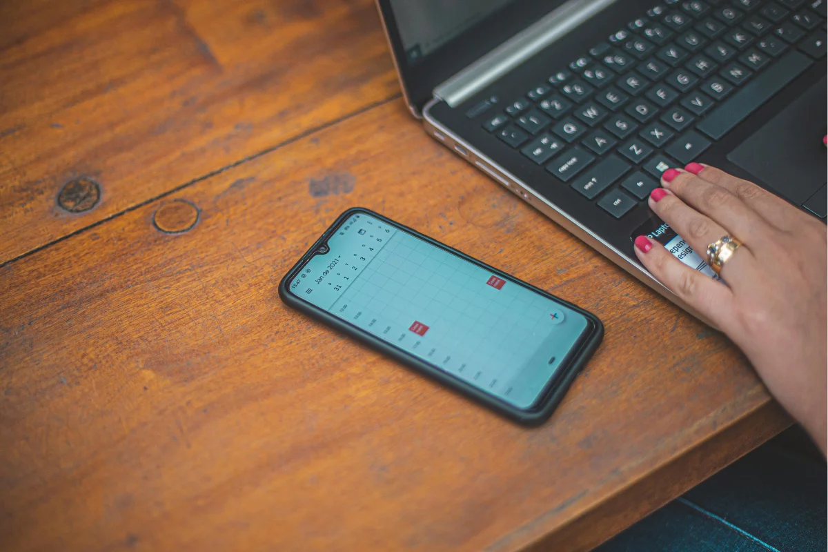 A smartphone displaying a calendar app sits on a wooden table next to a laptop keyboard, with a hand resting on the laptop